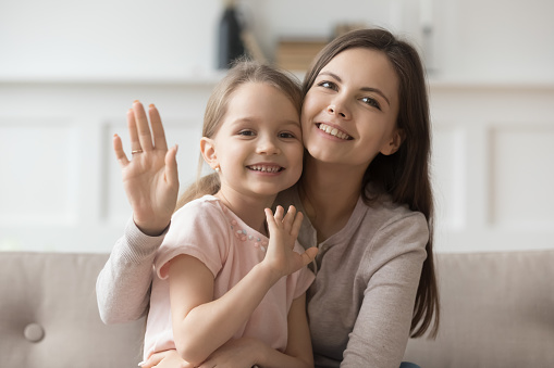 Portrait of smiling young mom hold hugging cute preschooler daughter look wave to camera, happy mother or nanny cuddle with little girl child shoot record video blog say hello to viewers