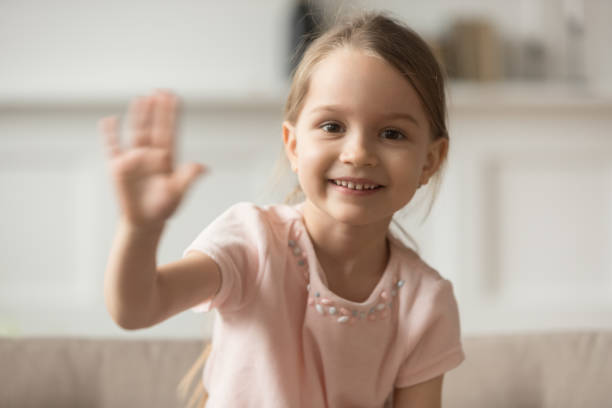 Little preschooler girl sit on couch waving looking at camera Funny preschooler girl sit on couch look at camera waving having webcam videochat conversation, smiling little child blogger shoot record video blog or vlog say hello to viewers subscribers kid waves stock pictures, royalty-free photos & images