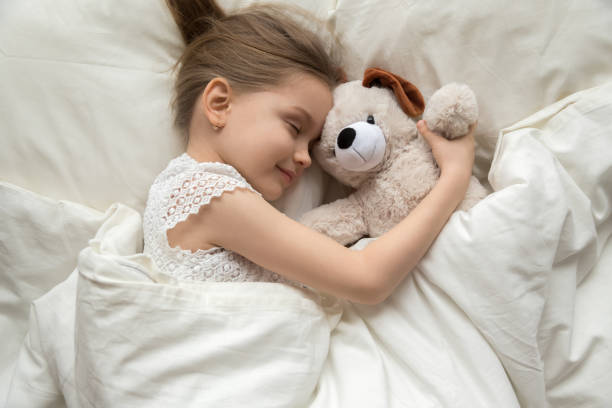 Preschooler girl hug teddy sleeping peacefully in cozy bed Smiling cute little girl hug teddy bear sleep peacefully in white bed relaxing on soft pillow, calm preschooler child enjoy daydream with plush toy taking nap under warm fluffy duvet at home fluffy blanket stock pictures, royalty-free photos & images