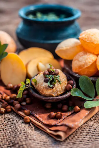 Famous Indian & Asian street food dish i.e. Panipuri snack in a clay bowl along with its flavored spicy water in another clay vessel. Entire consisting raw ingredients present on the surface.