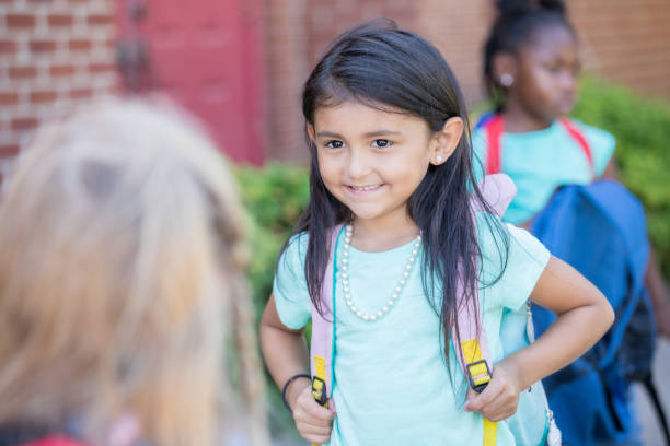 istock Elementary age Hispanic little girl smiles while talking to classmates outside school and wearing backpack 1163247766