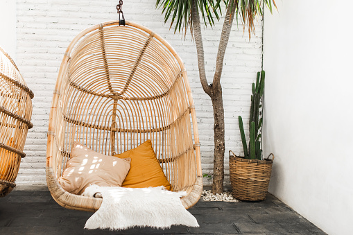 Wicker rattan hanging chair in loft cafe. Eco friendly furniture style and concept. Orange pillows and soft fur on chair. Hipster cafe in mexican style with cactus and palm.