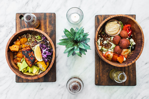 Flat lay of wooden bowls with fresh organic vegetarian food on white marble table. Falafel, hummus, avocado, vegetable salad, salmon steak. Olive oil on side. Healthy food concept.