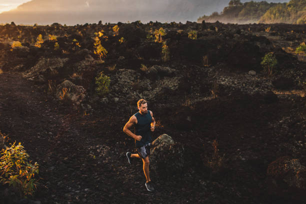 日の出時に山の中を走る若い男性アスリートトレイル。背景にバリ島の驚くべき黒い溶岩火山の風景。アドベンチャースポーツコンセプト。 - footpath single lane road sunrise landscape ストックフォトと画像