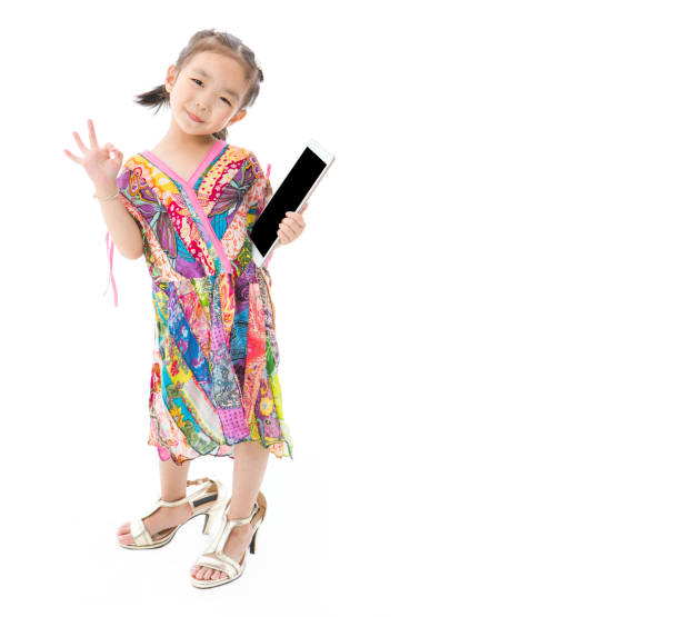 Portrait of cute little Asian girl in red eyeglasses wearing colorful dress, play with her mother’s high heels shoes, tablet in hands and making OK sign, smiling at camera on isolated white background