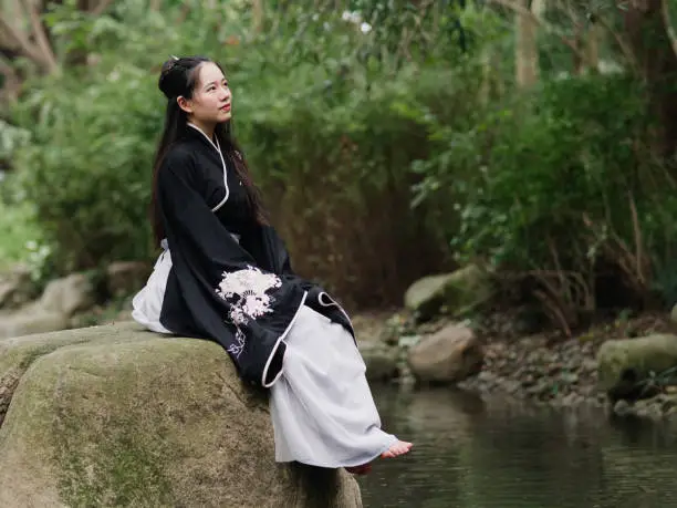 Beautiful Asian woman in black and white Chinese costume clothes hanfu sitting on big rock in middle of stream looking away in peaceful smile, traditional ancient Chinese beauty, time travel fiction.