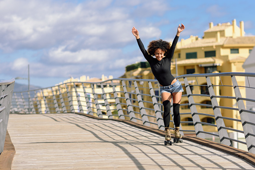 Black woman, afro hairstyle, on roller skates riding outdoors on urban bridge with open arms. Smiling young female rollerblading on sunny day. Beautiful clouds in the sky.