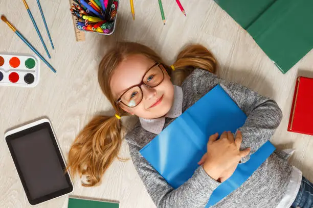 Photo of Cute little girl in glasses with a book lying on the floor. A child is surrounded by a book, tablet, paints, brushes, pencils.