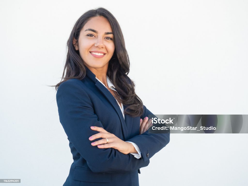 Smiling confident businesswoman posing with arms folded Smiling confident businesswoman posing with arms folded. Happy beautiful black haired young Latin woman in formal suit standing for camera over white studio background. Corporate portrait concept Businesswoman Stock Photo