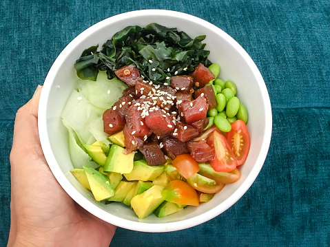 Close up tuna poke bowl with fresh sliced tomato, avocado, cucumber, tuna meat, edamame, cucumber, wakame, seaweed, sesame seed in a bowl