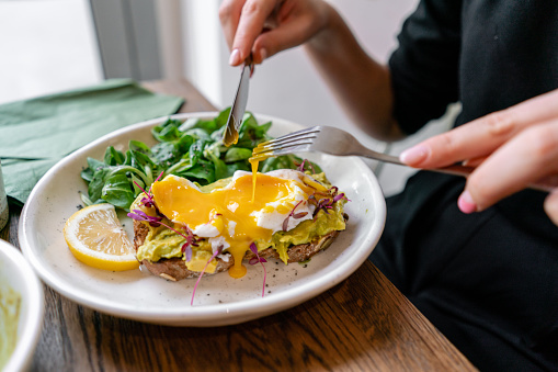 Young woman eat breakfast with knife and fork, Morning in cafe. Healthy breakfast with wholemeal bread toast with avocado, poached egg with green salad.