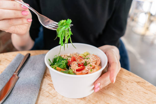 woman eating traditional hawaiian dish poke bowl with fork. shrimps with rice, radish,cucumber, tomato, sesame seeds and seaweeds. diet and useful fast food - poking prepared ahi tuna hawaiian culture imagens e fotografias de stock