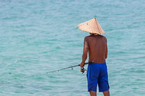 Koh Samui, Thailand - April 19, 2019: Local man with handmade bamboo hat fishing with a fish pole