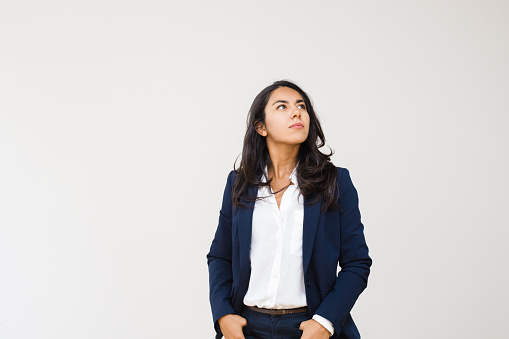 Confident businesswoman with hands in pockets looking up. Professional confident young businesswoman standing with hands in pockets isolated on grey background. Business concept