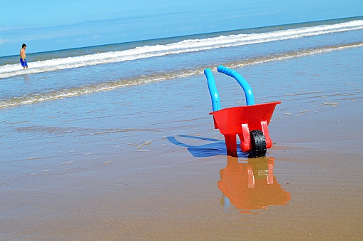 A red children's plastic wheelbarrow with blue handles and a black wheel on a sandy beach. Children's sand toy are on textured sand of a sandy beach in England. The object has a shadow as well as a beautiful reflection on the wet sand. A boy is standing far in the background