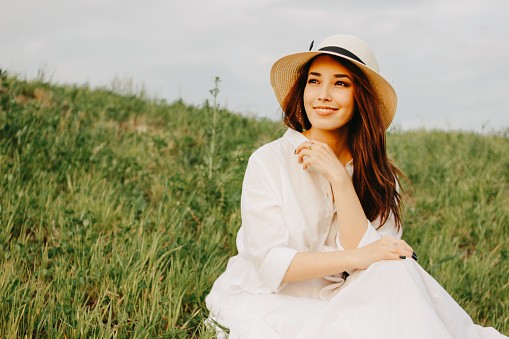 Charming carefree long hair asian girl in white clothes and straw hat enjoys life in field at sunset. Sensitivity to nature concept