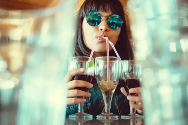 An indoor shoot of Asian/Indian young women in cool round sunglasses. She drinks three glasses of mock-tail drinks at once through straws in a restaurant. The image shot from a low angle through the gap between two blur glasses.