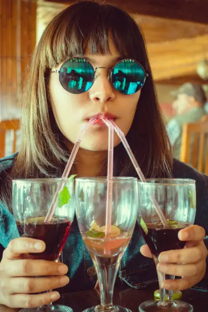 Indoor shoot of an Asian/Indian young women in cool round sunglasses drinks three glasses of mock-tail at once through straws in a restaurant.