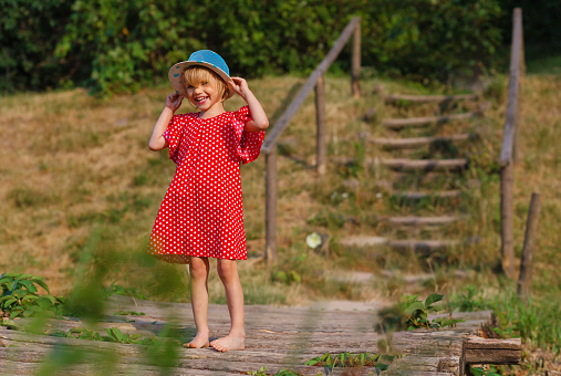 Little blonde hair and blue eyes kid girl in polka dot red dress walk barefoot on a wooden bridge enjoys a warm sunny day. Hiking and walking in nature. Leisure and outdoor activities.