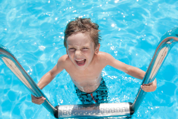 delightful small kid with wet hair and body standing on stainless handrail of blue water outdoor summer swimming pool during leisure travel - fashionable the human body short hair human head imagens e fotografias de stock