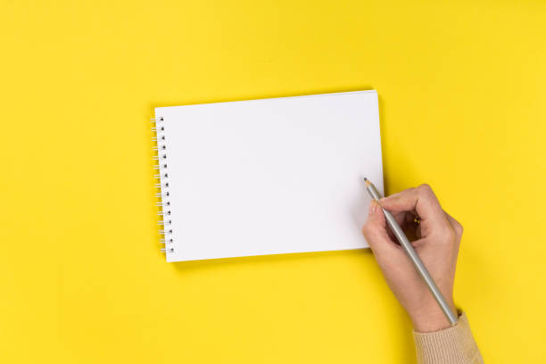 istock Woman's hands with perfect manicure holding pencil and  notepad as mockup for your design. Yellow background, flat lay 1163355751