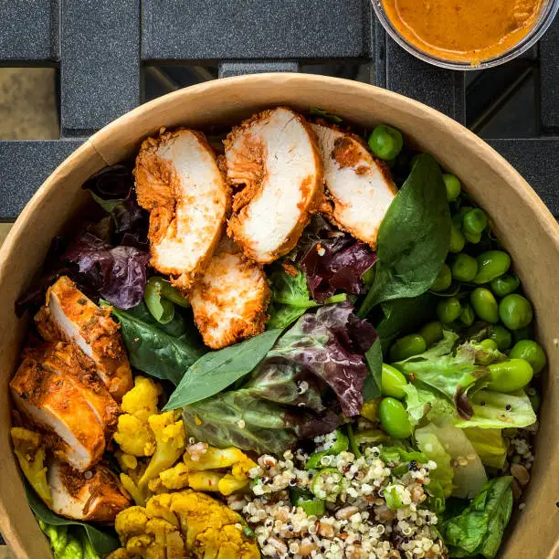 Photo of Overhead view of harissa chicken with mixed vegetable, grains, quinoa and harissa dressing on the side in a disposable box