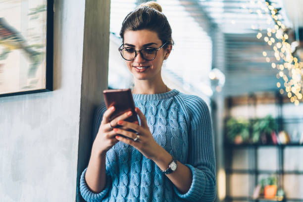Cheerful hipster blogger in eyeglasses reading funny comments under internet post holding modern smartphone in hands.Positive young woman laughing during online chat with friends on cellular