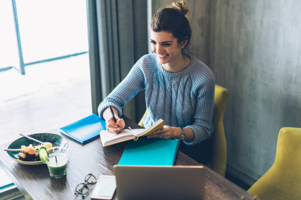 istock Positive young woman sitting at desktop with laptop computer and writing down checklist in notebook in cafe interior.Cheerful hipster blogger laughing while making notes of publication in notepad 1163365913