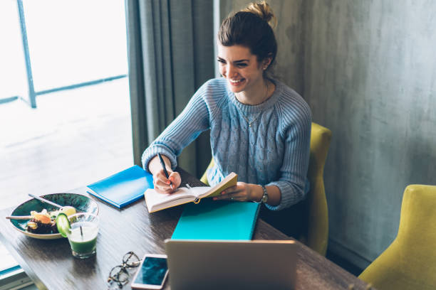 istock Cheerful hipster student writing down notes of design project in notebook sitting at laptop computer in coworking space.Positive young woman laughing and noting records of checklist in notepad 1163365923