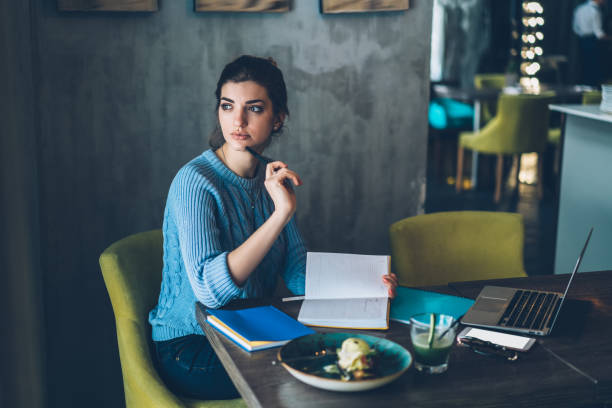 istock Contemplative young woman thinking on ideas for design project sitting at table with notepad and laptop computer in coffee shop.Dreamy hipster girl pondering on writing of creative essay 1163365944