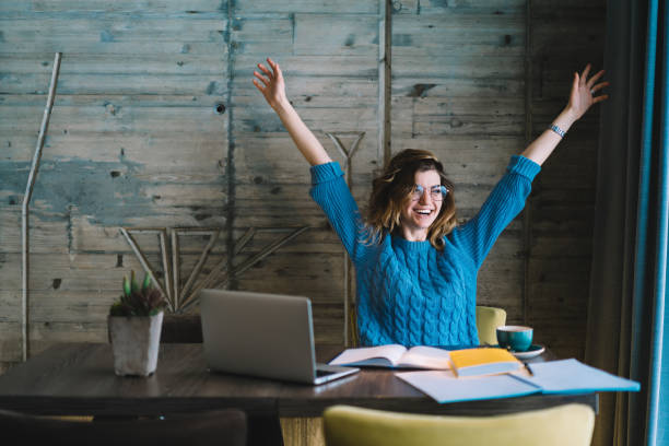 étudiant hipster heureux se réjouissant de la réussite du projet de formation tenant la main vers le haut. la jeune femme surheureuse a accompli la tâche d'étude pendant la préparation d'examen s'asseyant au bureau avec l'ordinateur portatif et le ma - résultat dexamen photos et images de collection