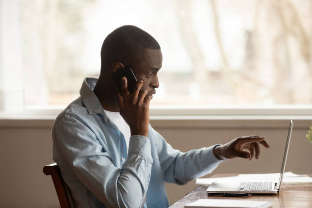African guy looking at computer screen talk on phone African 30s man sitting at table looking pointing at computer screen discuss application talking on mobile phone working at home solve business issues distantly, self-employed male speak with client melancholy computer support specialist stock pictures, royalty-free photos & images