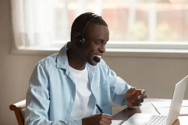 Photo of African guy wear headset making notes studying online using computer