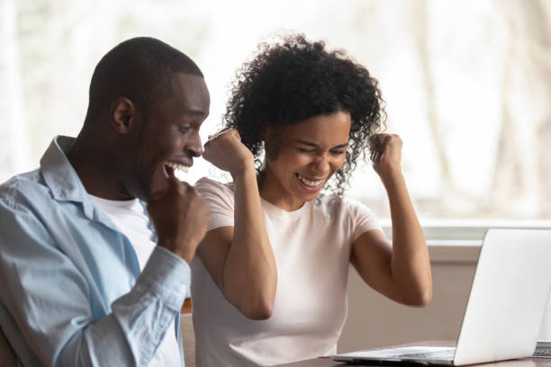 istock Overjoyed african couple celebrate online victory sit at table 1163367356