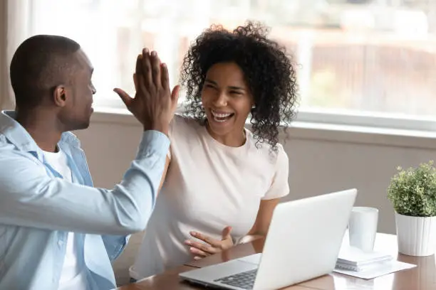 Happy african husband biracial wife couple sit at table near computer make fists yes gesture celebrating victory feels overjoyed unbelievable luck online lottery win betting, mortgage approval concept