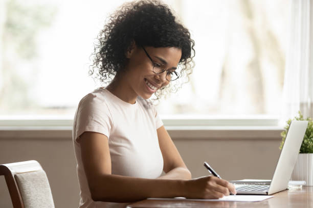 African woman in glasses sitting near computer holding pen writing Satisfied black woman housewife in glasses sitting at table near laptop holding pen writing planning family budget making notes, businesswoman working at home, girl studying online noting information woman examining stock pictures, royalty-free photos & images