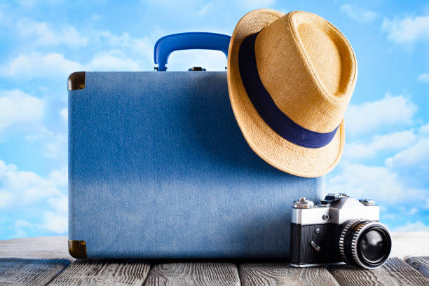 Blue suitcase, summer hat and vintage photo camera on wooden table and beach sand background stock photo