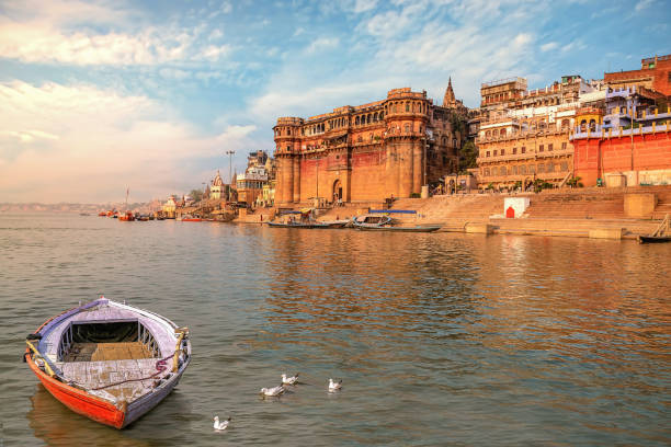 Varanasi ancient city architecture at sunset with view of boat on river Ganges Ancient Varanasi India city architecture with view of Ganges river ghat at sunset. Wooden boat seen on river Ganga with migratory birds varanasi photos stock pictures, royalty-free photos & images