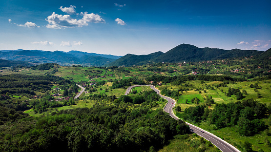 Color panoramic image depicting an aerial view of a green rural landscape in the Transylvania region of Romania. The area is heavily forested with hills in the distance and blue sky with cloudscape. A winding road runs directly through the image, with tiny cars seen from above. Room for copy space.