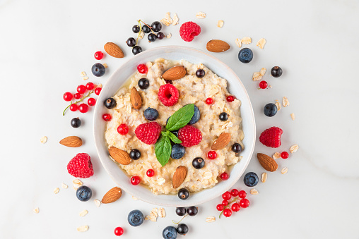 bowl of oatmeal porridge with fresh berries, nuts and mint for healthy diet breakfast on white marble table. top view