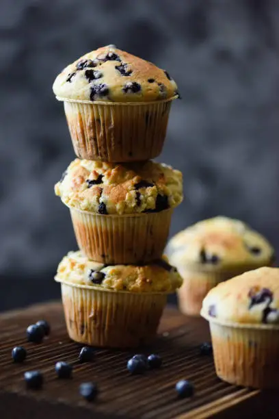 Unstable pile of freshly baked blueberry muffins on oak cutting board on dark background. Low key still life with natural lighting side view copy space