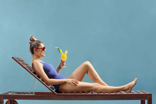 Full length shot of a young woman relaxing on a sunbed with a cocktail isolated on blue background