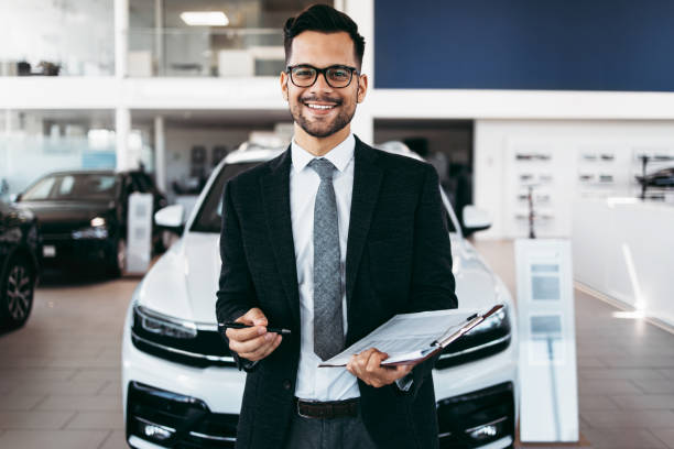 Car dealer standing and posing in showroom Good looking, cheerful and friendly salesman poses in a car salon or showroom and looks at camera. car salesperson photos stock pictures, royalty-free photos & images