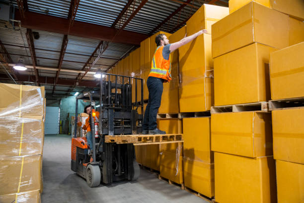 An industrial warehouse workplace safety topic. A dangerous situation as a worker is lifted-up by a forklift to reach higher placed merchandise. stock photo