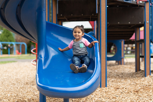 A cute young Native American girl comes to the bottom of a slide on the playground. She has a big smile for the camera.