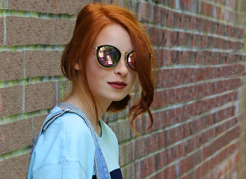 Stock photo of pretty moody, sad teenage girl youth with red hair looking at camera, smiling lost in thought by brick wall, redhead wearing blue denim dungarees, sunglasses, makeup, dark red lipstick, teenager with ginger hair tied back in curls