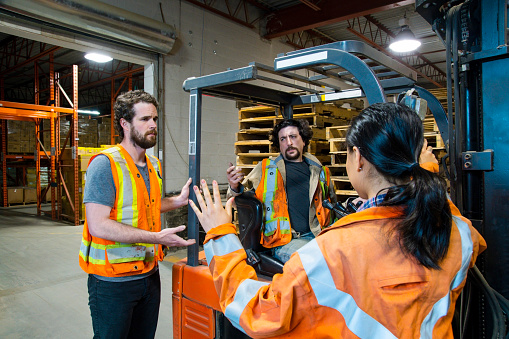 An industrial warehouse workplace safety topic. A manager discusses an issue with a forklift driver and a coworker.