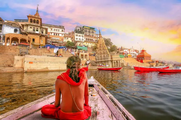 Sadhu baba sitting on a wooden boat overlooking ancient Varanasi city architecture with Ganges river ghat at sunset.