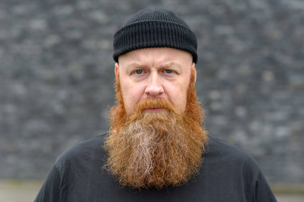 Intense man scowling at the camera Intense man with bushy red beard wearing a beanie hat scowling at the camera with a penetrating stare over grey bushy stock pictures, royalty-free photos & images
