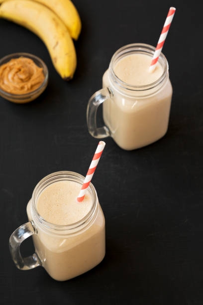 Mason glass jar mugs filled with peanut butter banana smoothie on a black background, side view. Close-up.